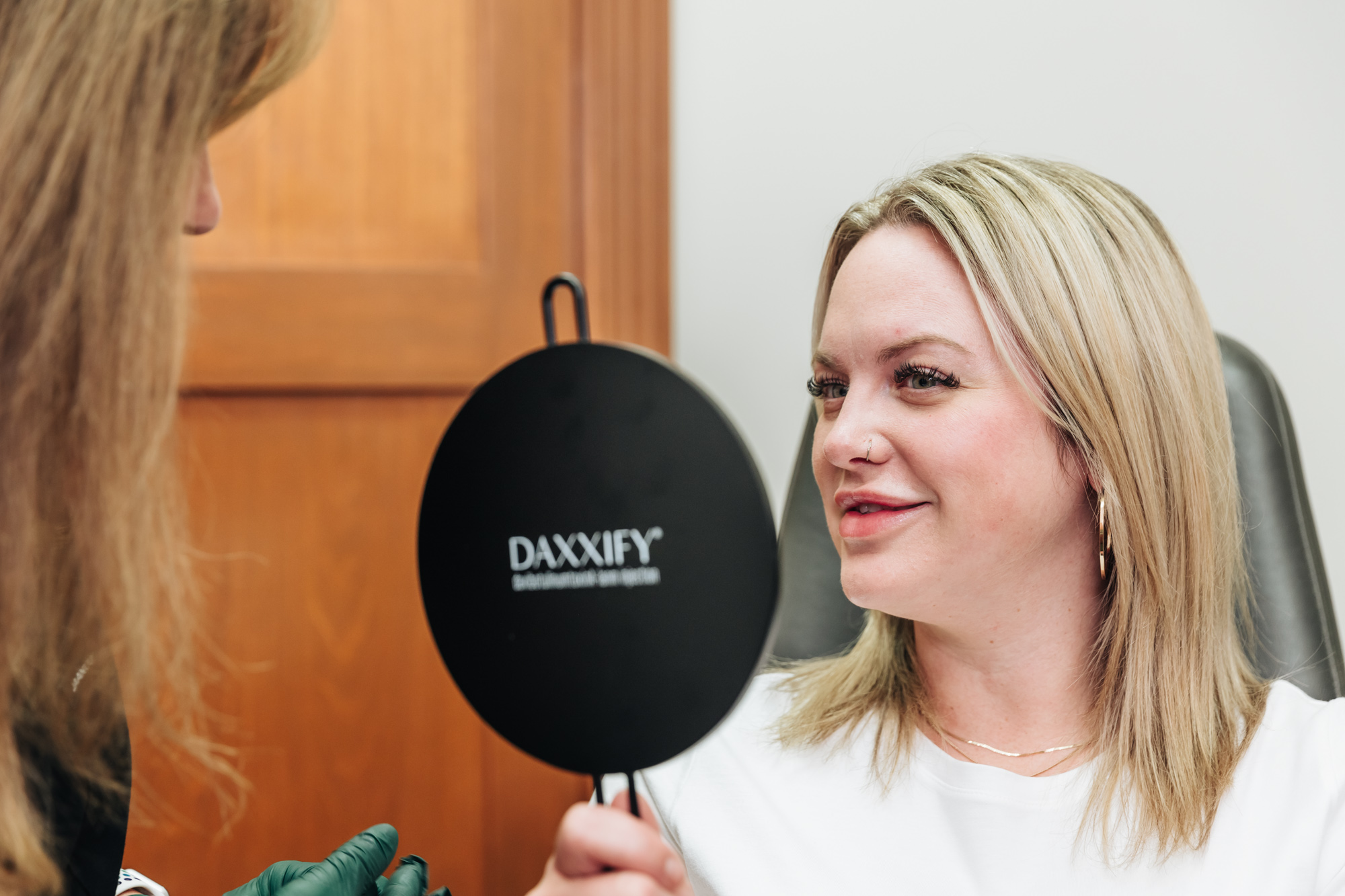 a woman hols up a mirror and examines her face as she learns about Acne Scar Treatment in Portage